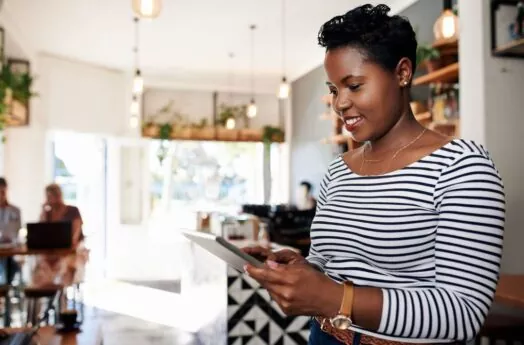 Uma mulher negra está em uma casa bem decorada segurando um tablet. Ela está sorrindo e usa uma camisa com listras horizontais pretas e brancas, em seu braço está um relógio dourado.